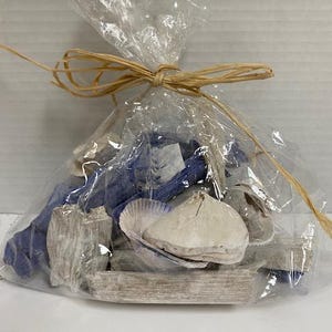 May include: A clear cellophane bag tied with raffia, filled with decorative seashells, driftwood, and blue fabric pieces. The bag contains various shapes and sizes of natural elements, creating a coastal theme.