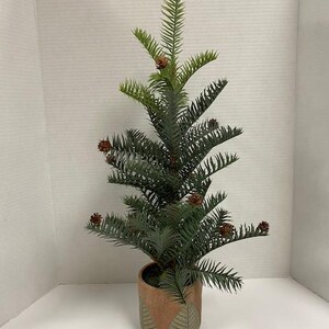 May include: A small, artificial evergreen tree in a decorative pot. The tree has dark green, needle-like leaves and small brown pine cones. The pot is a light brown color with a geometric design in shades of green and brown.