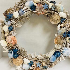 May include: A circular wreath made of natural twigs, adorned with seashells in shades of white, tan, and blue. Small blue fabric flowers and dried foliage are interspersed throughout the design, creating a coastal theme.