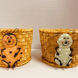 May include: Two decorative ceramic baskets with insect designs. One basket features a ladybug design with orange and black accents. The other basket has a bee design with green and brown accents. Both baskets have a woven texture.