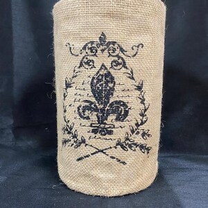May include: A cylindrical burlap vase with a black fleur-de-lis design. The design includes a floral wreath, script, and crossed stems. The vase is tan and has a rough texture.