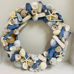 May include: A circular wreath made of seashells, driftwood, and blue-painted wood pieces. The wreath features various types of seashells, including starfish, and is arranged in a decorative pattern. The color palette includes natural tones, blues, and whites.