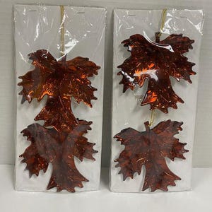 May include: Two sets of two hanging ornaments shaped like maple leaves, each made of a shiny, translucent dark amber-colored material. The leaves feature detailed vein patterns and are attached to gold-colored strings, packaged in clear plastic.