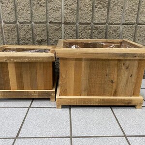 May include: Two rectangular wooden planters of different sizes, constructed from light brown vertical wood planks with a thick top rim and a raised base. Each planter features a clear plastic liner visible inside the open top compartment.