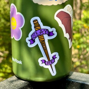 May include: A green water bottle decorated with stickers. A sticker shows a dagger with a purple banner reading "SLAY ME". Other stickers include a purple flower with a yellow smiley face and a bat.