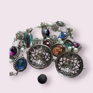 May include: A silver necklace with multiple caged pendants, each containing a different coloured gemstone. The necklace is adorned with small white beads and features a variety of colourful gemstones, including blue, green, red, and purple.