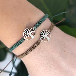 Könnte beinhalten: Zwei Makramee-Armbänder mit einem Lebensbaum-Anhänger. Eines ist dunkelgrün, das andere braun. Der Lebensbaum-Anhänger ist silberfarben. Die Armbänder befinden sich an einem Handgelenk.