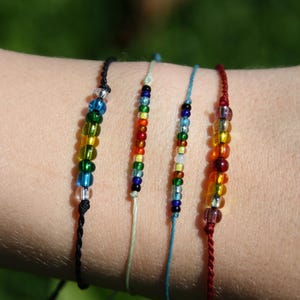 Può includere: Cinque braccialetti colorati con perline su un polso. I braccialetti sono realizzati con piccole perline di vetro in tonalità di verde, blu, giallo, rosso e arancione. I braccialetti sono legati con sottili cordini in nero, bianco, blu e rosso.