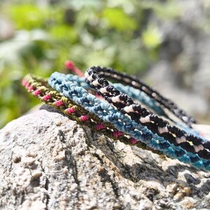 Könnte beinhalten: Nahaufnahme von mehreren handgefertigten Makramee-Armbändern in verschiedenen Farben, darunter Grün, Blau, Schwarz und Rosa. Die Armbänder liegen auf einer strukturierten Felsfläche mit verschwommenem grünem Hintergrund.
