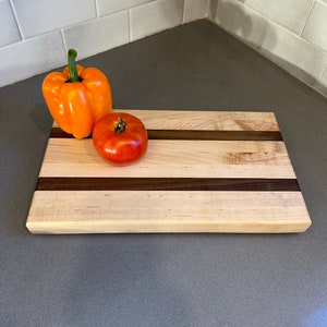 May include: A rectangular wooden cutting board with two dark brown stripes. An orange bell pepper and a red tomato rest on the board. The cutting board is made of light-colored wood.