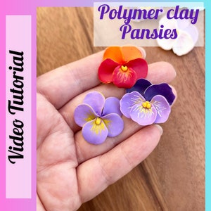 May include: Four polymer clay pansies in different colors: purple, red, orange, and white. The pansies are in a person's hand. The image is titled "Polymer clay Pansies" and "Video Tutorial".