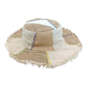 Könnte beinhalten: Ein Patchwork-Eimerhut mit ausgefranstem Rand. Der Hut hat beige, hellbraune und weiße Stoffbahnen mit farbigen Nähten. Das Oberteil hat einen weißen Streifen und ein blaues Zickzack-Muster.
