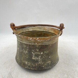 May include: A weathered, cylindrical metal bucket featuring a dark, oxidised patina with visible surface wear and discolouration. The vessel includes a thick, curved metal handle attached to two side loops, with a wide, open top and a rounded base.