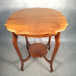 May include: A medium-brown wooden side table featuring a scalloped edge tabletop and a matching smaller scalloped shelf positioned between four curved legs. The wood grain is visible across the surfaces, and the structure is elevated on four legs.