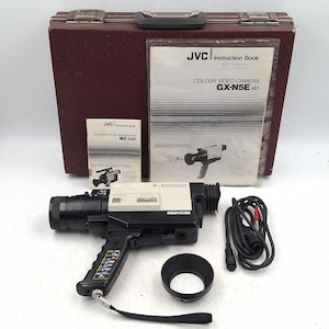 May include: Vintage JVC Colour Video Camera GX-N5E (c) with instruction manuals and a carrying case. The camera is black and white with a black lens and handle. Includes a lens hood and cables.