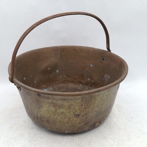 May include: A round, weathered copper pot featuring a wide, open top and a curved, dark metal handle attached to the sides. The exterior surface displays a tarnished, multi-toned metallic finish with visible signs of age, oxidation, and wear.
