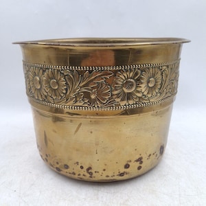May include: A polished brass planter featuring a wide, embossed band around the upper section. The decorative band displays a repeating pattern of daisy-like flowers and leafy foliage, framed by two rows of small, raised beaded detailing.