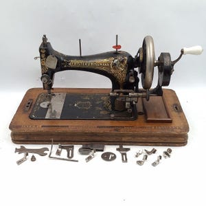 May include: An antique Frister & Rossmann sewing machine with a wooden base and various attachments. The machine is black with gold detailing and a silver hand wheel. Additional tools are arranged in front of the machine.