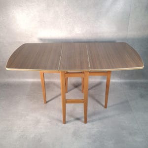 May include: A mid-century drop-leaf table with a brown wood grain surface and light brown legs. The table has a rectangular shape with rounded corners and a light-colored trim. The table is approximately 122 cm long when fully extended.