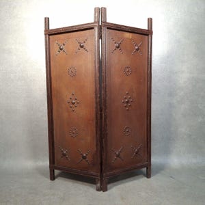 May include: A three-panel folding screen with a dark brown wooden frame and panels. The panels are a lighter brown and decorated with metal studs in circular and cross patterns. The screen is freestanding and has a simple, rustic design.