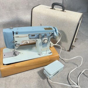 May include: A light blue Westminster sewing machine mounted on a wooden base, accompanied by a matching light blue foot pedal and a beige carrying case with a dark handle. The machine features various silver dials, levers, and a power cord.