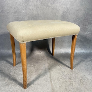 May include: A rectangular stool featuring a cream-coloured upholstered seat with a subtle diamond pattern and tufted buttons. The seat is finished with a decorative braided trim along the edge and is supported by four straight, light-toned wooden legs.
