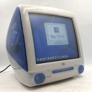 May include: A vintage iMac G3 desktop computer featuring a translucent blue and white plastic casing. The screen displays the Mac OS 9.1 startup interface with a loading bar. The front includes integrated speakers and a slot-loading optical drive.