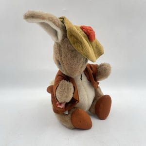 May include: A soft toy rabbit, dressed in a brown jacket and a yellow corduroy hat with a red detail. The rabbit has long ears and is in a seated position. The toy is made of soft, plush material.