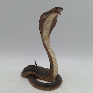 May include: A brown and cream-coloured cobra figurine in an upright, hooded striking pose. The snake features a textured, scaly surface with a dark brown coiled base, a lighter brown hood, and a cream-coloured underside with horizontal ribbing.