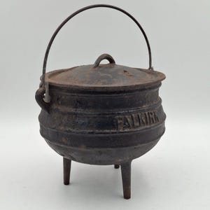 May include: A black cast iron pot with three legs, a matching lid, and a wire bail handle. The word FALKIRK is embossed on the side of the rounded body. The surface shows a weathered, dark texture consistent with heavy metal cookware construction.