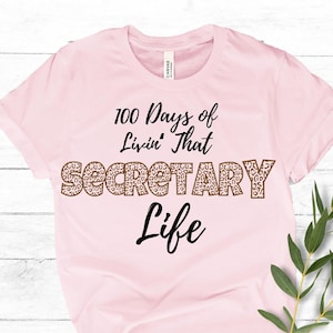 May include: A light pink t-shirt with the text "100 Days of Livin' That SECRETARY Life" in black and leopard print. The shirt is made of a soft material and has short sleeves. The shirt is displayed on a white wooden surface.