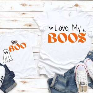 May include: White t-shirts with Halloween graphics. One says "little BOO" with a ghost, the other "Love My BOOS" with bats. They're styled with blue jeans and white trainers. The items are on a wooden surface.
