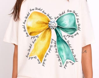 Puede incluir: Una camiseta blanca con una ilustración de acuarela de un lazo amarillo y verde. El lazo está rodeado por el texto "House Divided" repetido varias veces.