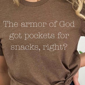 Puede incluir: Camiseta marrón con el texto "The armor of God got pockets for snacks, right?" en blanco. La camiseta tiene cuello redondo y está anudada a la cintura. La camiseta está hecha de un material suave.