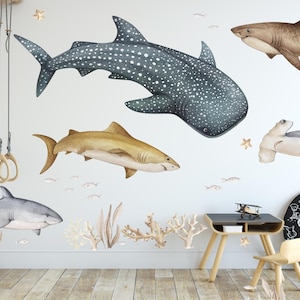 May include: A white wall with a variety of watercolor shark decals. The decals include a whale shark, a hammerhead shark, a brown shark, and a grey shark. The decals are arranged in a whimsical pattern on the wall. The text "Home of the Cool Kids" is written on a black chalkboard.