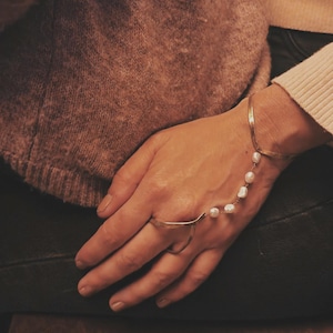 Op de afbeelding: Een goudkleurige handkettingarmband met een ring en een armband verbonden door een ketting van witte parels. De armband zit om de hand van een persoon, met een bruine trui op de achtergrond.