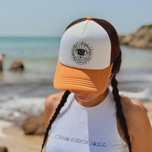 Puede incluir: Gorra de camionero con un panel frontal blanco con un diseño de ojo, una parte trasera de malla marrón y una visera naranja. La gorra tiene un botón naranja en la parte superior. El texto "PIENSA SIENTE HAZLO" es visible en la camiseta sin mangas blanca.