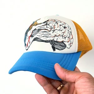 Puede incluir: Gorra de camionero con visera azul y panel frontal blanco con una ilustración detallada de un cerebro. La parte trasera de malla es de color amarillo dorado. La gorra está sujeta por una mano.