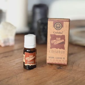 Puede incluir: Una pequeña botella marrón de aceite esencial con tapón blanco y etiqueta que dice "cinnamon". Junto a ella, una caja a juego con la marca "Goloka" y la palabra "cinnamon" impresa. El fondo está borroso.