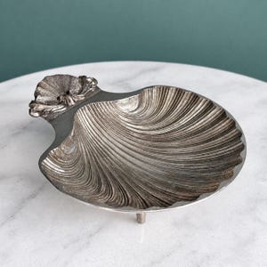 May include: A silver-toned metal dish shaped like a scallop shell with a textured, ribbed surface and a decorative hinge detail at the top. The dish is supported by small feet, featuring a metallic finish with dark highlights in the grooves.