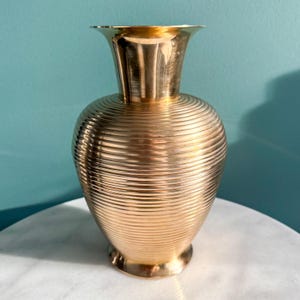 May include: A gold-coloured vase with a wide, fluted top and a rounded body. The body features horizontal, ridged detailing. The vase sits on a small, circular base. The background is a solid teal colour.