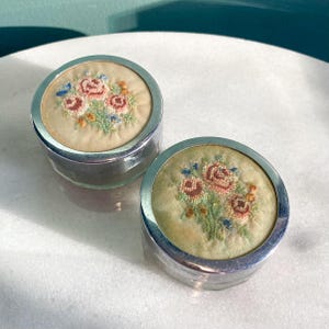 May include: Two small, round, silver-coloured containers with embroidered floral designs on the lids. The embroidery features red roses, blue accents, and green leaves on a beige background. Likely vintage or antique items.