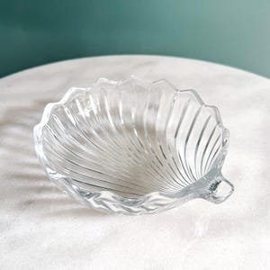 May include: A clear glass dish shaped like a scallop shell, featuring a scalloped edge and radiating vertical ridges that extend from the base to the rim. The bowl has a smooth, transparent finish and a distinct hinge-like detail at the bottom.
