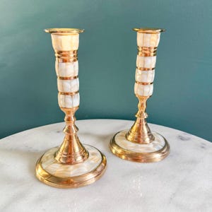 May include: Two vintage brass candlesticks with a decorative design. The candlesticks have a white and gold segmented body and a round base. The candle holders are displayed on a white marble surface against a teal background.