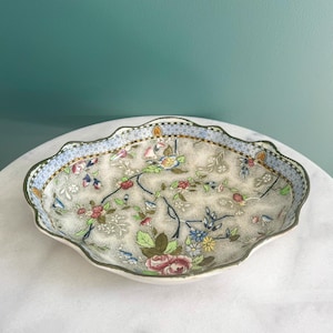 May include: An oval ceramic serving dish with a scalloped edge. The dish has a floral pattern with pink, blue, and yellow flowers on a cream background. A blue border with a geometric design frames the floral pattern.