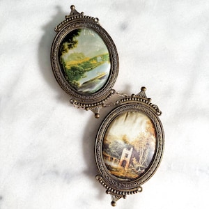 May include: Two oval-shaped brass frames connected by a small metal chain. Each frame features an ornate, textured border and contains a landscape painting. The top painting depicts a river and hills, while the bottom painting shows a stone building.