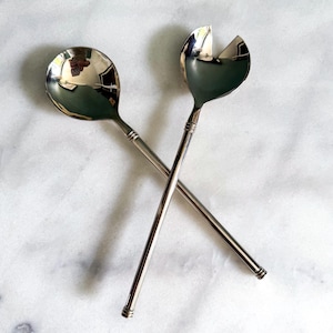 May include: Two polished silver salad servers, a spoon and a fork, are crossed against a white marble surface. The spoon has a rounded bowl, and the fork has a curved design. The handles are long and slender, with a decorative detail.