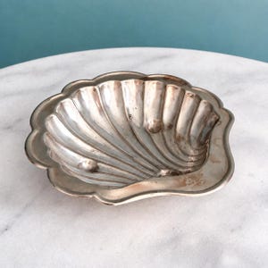 May include: A silver-toned metal dish shaped like a scallop shell with a scalloped edge and a series of radiating ridges. The interior surface features a smooth, reflective finish with subtle signs of tarnish and three small rounded feet.
