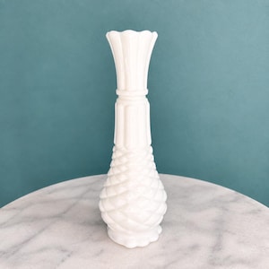 May include: A tall, opaque white milk glass vase featuring a flared, scalloped rim, a slender neck with vertical fluting, and a bulbous base decorated with a raised, diamond-shaped pineapple pattern that extends from the bottom to the midsection.