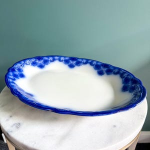 May include: An oval ceramic serving platter featuring a white centre and a wide, scalloped rim decorated with a deep blue floral pattern. The edges of the dish are embossed with intricate scrollwork details that frame the central white basin.